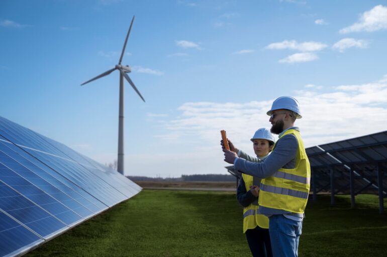 Painéis solares e hélices de energia eólica instalados em um campo com técnicos fazendo medição, representando a transição energética.