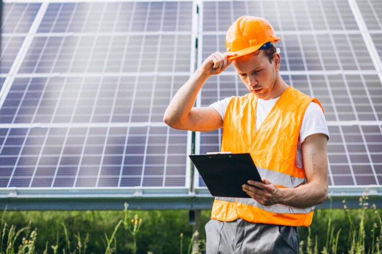 Pessoa com EPI e tablet na mão em frente a painéis solares, realizando análise de desempenho da energia solar.