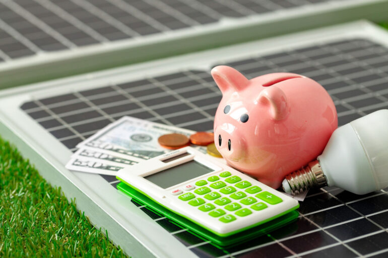 Dinheiro, calculadora, cofrinho e lâmpada sobre um painel solar, representando os impactos econômicos da energia solar.